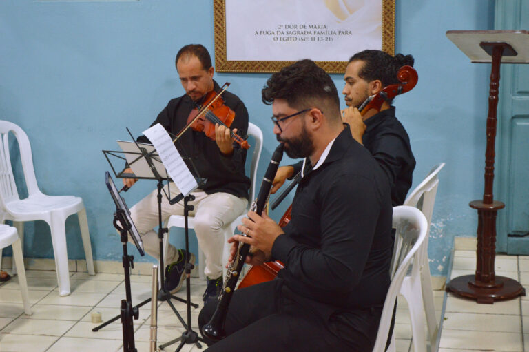 Professores da Vila se apresentam na Capela Nossa Senhora das Graças, na missa em homenagem aos 106 anos do Padre Ágio. São três professores: Nielson, à esquerda do leitor, tocando violino; Leone no meio, tocando violoncelo. Thomas na ponta direita do leitor, tocando clarinete.