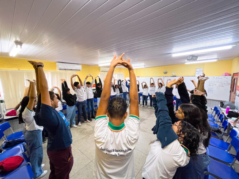 Na foto, o professor Vinícius e os alunos estão com os braços erguidos, se alongando durante a oficina de percussão corporal.