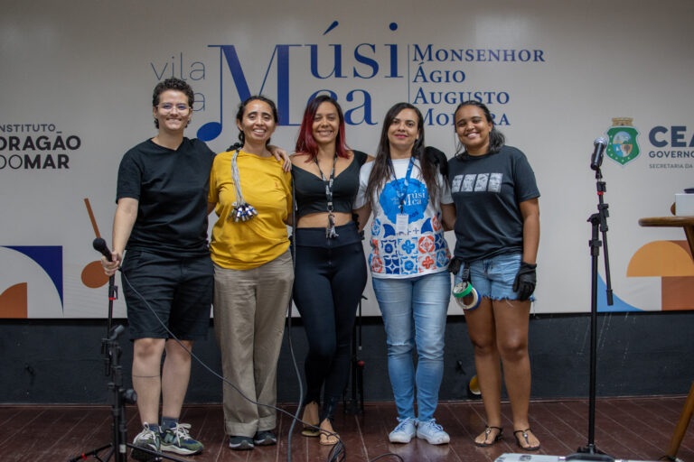 Na foto, cinco mulheres estão lado a lado, sorrindo e posando para a câmera em um palco da Vila da Música Monsenhor Ágio Augusto Moreira. A parede ao fundo exibe logotipos da instituição e do Governo do Estado do Ceará. Da esquerda para a direita: Ravena Monte, vestindo preto e segurando um microfone, foi a responsável por ministrar a Oficina de Roadie para Mulheres - Módulo II. Ao seu lado, uma mulher de blusa amarela e calça bege, com um feixe de cabos pendurado no ombro. No centro, uma mulher de blusa preta e calça justa posa sorridente. A quarta participante veste uma camisa branca e azul com a logo da Vila da Música. Por fim, a última mulher à direita veste uma camiseta preta e shorts jeans, segurando fita adesiva e vestindo luvas, itens comuns no trabalho de roadie. A imagem representa a importância da capacitação e da inclusão de mulheres no setor técnico da música.