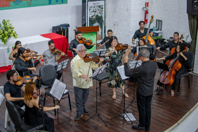 A imagem retrata uma apresentação musical com uma orquestra de câmara no palco de um auditório. Músicos tocam violinos, violas, violoncelos e contrabaixo, seguindo a regência de um maestro. Em destaque, dois solistas violinistas estão ao centro, um homem de camisa amarela e uma mulher com vestido estampado, ambos concentrados na execução da peça. O ambiente sugere um concerto formal, com partituras em estantes e uma mesa decorada ao fundo.