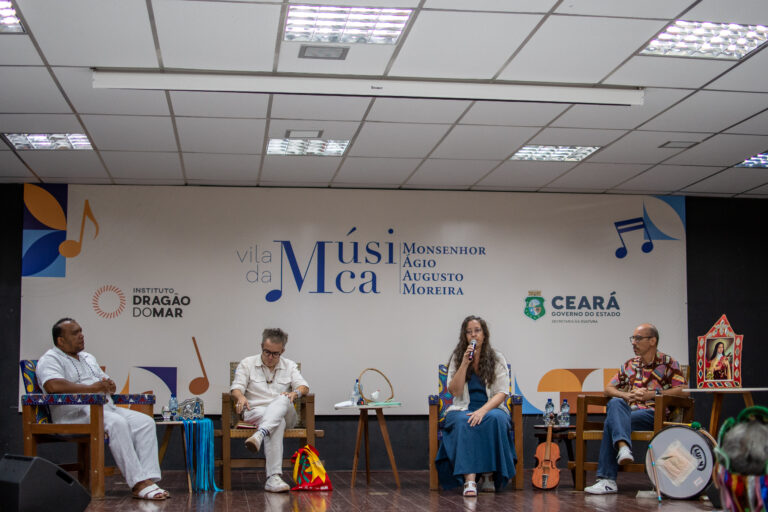 Integrantes da mesa-redonda “Acervos e Cartografias Sonoras”. Da esquerda para a direita: Mestre Nando Guerreiro, um homem negro, de roupa e sandálias brancas; Fabiano Piúba, homem branco também de roupas brancas; professora Letícia Bertelli, uma mulher branca, de cabelos castanhos longos, usando um vestido azul e casaquinho branco e calçando um sapato branco; por fim, o professor Márcio Mattos, usando camisa estampada colorida, em tons de rosa e vermelho, calça jeans e sapatos brancos com detalhes em preto. Todos estão sentados em cadeiras de couro, em cima do palco, onde há um banner da Vila da Música ao fundo.