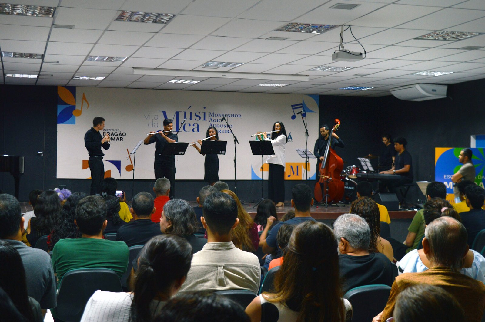 A imagem mostra uma apresentação musical na Vila da Música Monsenhor Ágio Augusto Moreira. No palco, há um grupo de músicos tocando instrumentos como flauta, contrabaixo, teclado e bateria, enquanto o público assiste atentamente. O ambiente é uma sala de concerto com iluminação de teto, e o fundo do palco apresenta um painel com o nome da instituição e elementos gráficos relacionados à música.