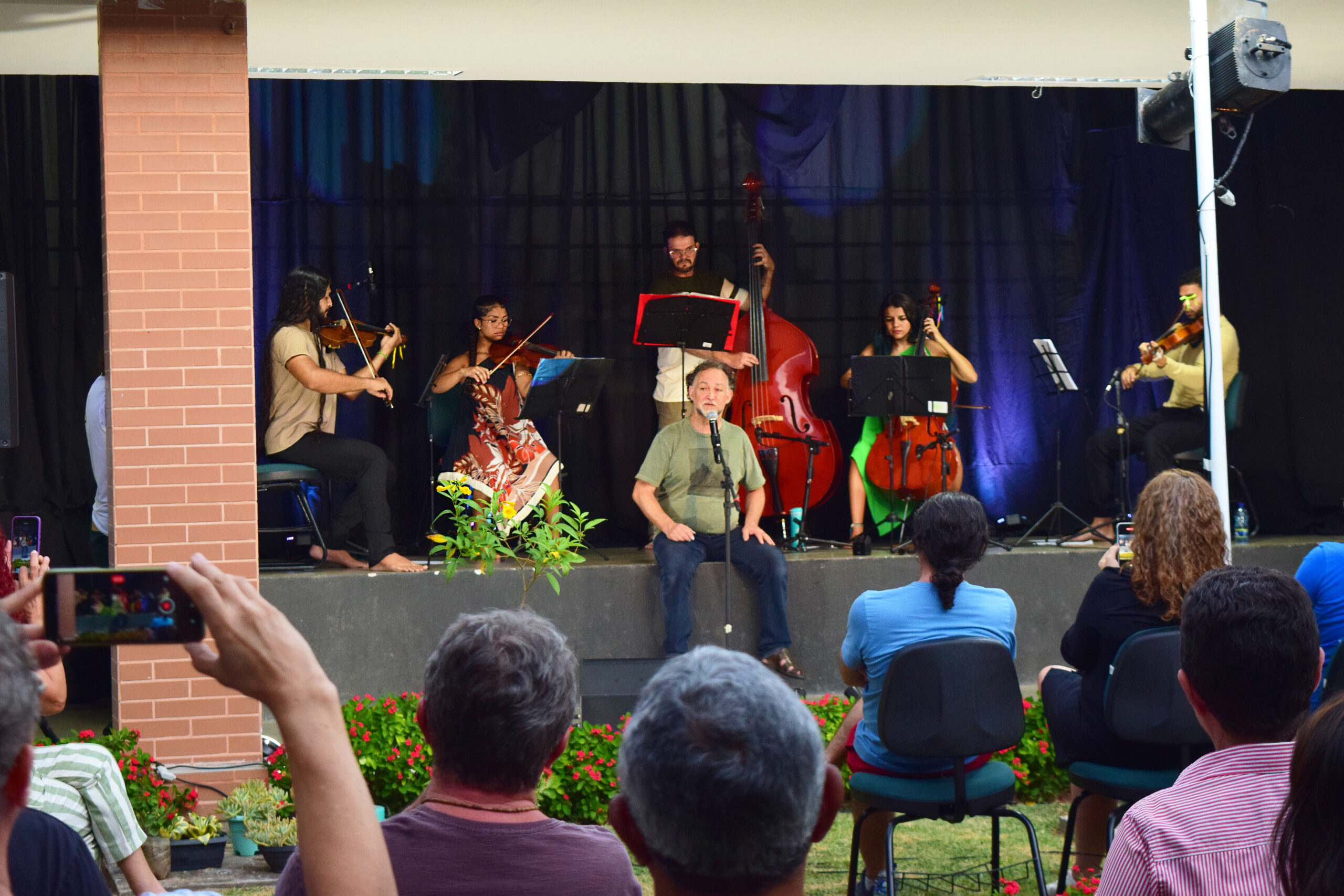 Fotografia do espetáculo musical ao ar livre, Sarau "Sons da Vila". No centro do palco, Claudio Nucci, de camiseta verde e calça jeans está sentado, cantando ao microfone. Atrás dele, seis instrumentistas tocam em semicírculo: dois violinistas, uma violista, uma violoncelista, um contrabaixista e outro músico de cordas. Os músicos são do grupo Ybirá. O cenário tem cortinas escuras e iluminação azulada. Em frente ao palco, pessoas do público assistem sentadas em cadeiras, algumas registrando a apresentação com celulares. Há plantas e flores vermelhas na área em frente ao palco.
