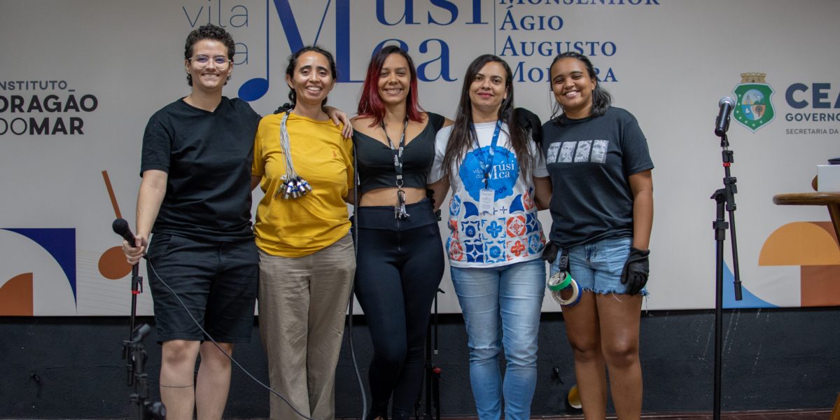 Na foto, cinco mulheres estão lado a lado, sorrindo e posando para a câmera em um palco da Vila da Música Monsenhor Ágio Augusto Moreira. A parede ao fundo exibe logotipos da instituição e do Governo do Estado do Ceará. Da esquerda para a direita: Ravena Monte, vestindo preto e segurando um microfone, foi a responsável por ministrar a Oficina de Roadie para Mulheres - Módulo II. Ao seu lado, uma mulher de blusa amarela e calça bege, com um feixe de cabos pendurado no ombro. No centro, uma mulher de blusa preta e calça justa posa sorridente. A quarta participante veste uma camisa branca e azul com a logo da Vila da Música. Por fim, a última mulher à direita veste uma camiseta preta e shorts jeans, segurando fita adesiva e vestindo luvas, itens comuns no trabalho de roadie. A imagem representa a importância da capacitação e da inclusão de mulheres no setor técnico da música.