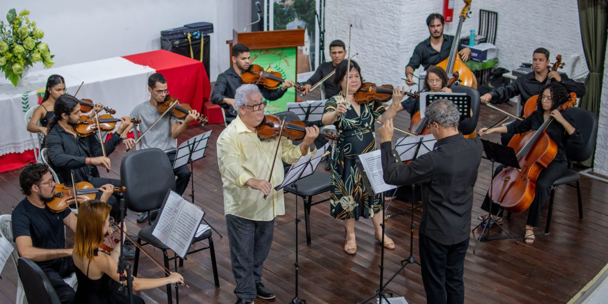 A imagem retrata uma apresentação musical com uma orquestra de câmara no palco de um auditório. Músicos tocam violinos, violas, violoncelos e contrabaixo, seguindo a regência de um maestro. Em destaque, dois solistas violinistas estão ao centro, um homem de camisa amarela e uma mulher com vestido estampado, ambos concentrados na execução da peça. O ambiente sugere um concerto formal, com partituras em estantes e uma mesa decorada ao fundo.