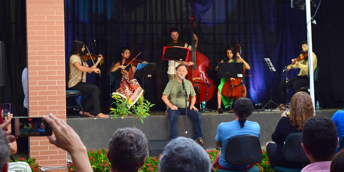 Fotografia do espetáculo musical ao ar livre, Sarau "Sons da Vila". No centro do palco, Claudio Nucci, de camiseta verde e calça jeans está sentado, cantando ao microfone. Atrás dele, seis instrumentistas tocam em semicírculo: dois violinistas, uma violista, uma violoncelista, um contrabaixista e outro músico de cordas. Os músicos são do grupo Ybirá. O cenário tem cortinas escuras e iluminação azulada. Em frente ao palco, pessoas do público assistem sentadas em cadeiras, algumas registrando a apresentação com celulares. Há plantas e flores vermelhas na área em frente ao palco.