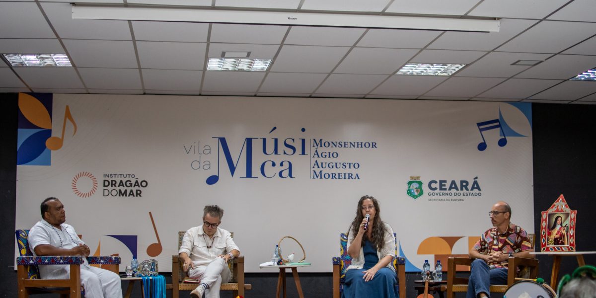Integrantes da mesa-redonda “Acervos e Cartografias Sonoras”. Da esquerda para a direita: Mestre Nando Guerreiro, um homem negro, de roupa e sandálias brancas; Fabiano Piúba, homem branco também de roupas brancas; professora Letícia Bertelli, uma mulher branca, de cabelos castanhos longos, usando um vestido azul e casaquinho branco e calçando um sapato branco; por fim, o professor Márcio Mattos, usando camisa estampada colorida, em tons de rosa e vermelho, calça jeans e sapatos brancos com detalhes em preto. Todos estão sentados em cadeiras de couro, em cima do palco, onde há um banner da Vila da Música ao fundo.