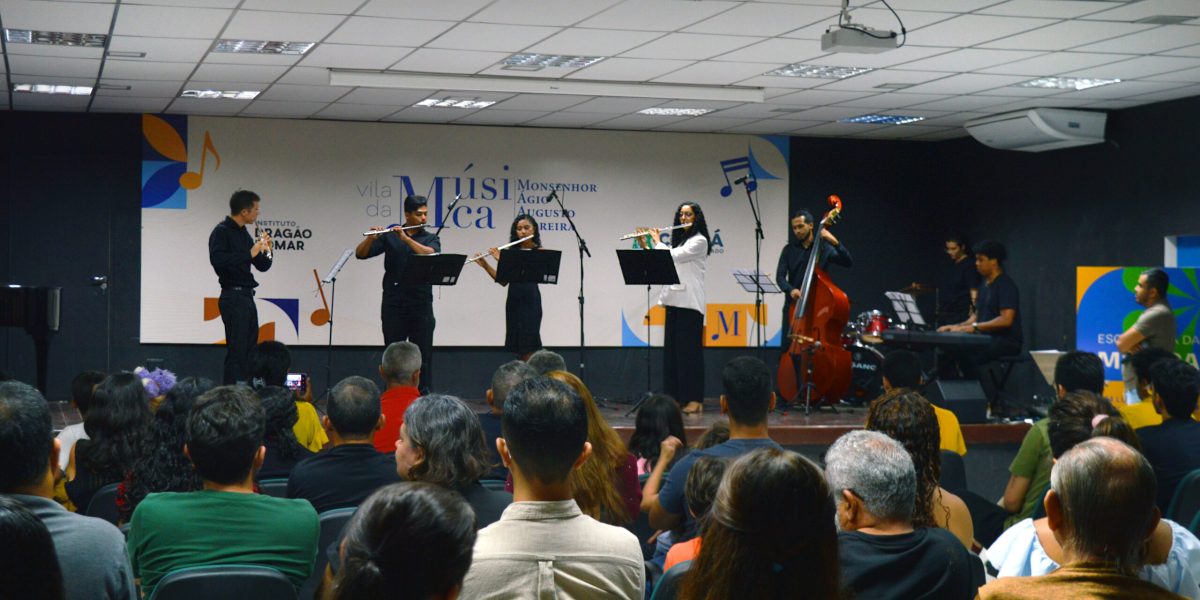 A imagem mostra uma apresentação musical na Vila da Música Monsenhor Ágio Augusto Moreira. No palco, há um grupo de músicos tocando instrumentos como flauta, contrabaixo, teclado e bateria, enquanto o público assiste atentamente. O ambiente é uma sala de concerto com iluminação de teto, e o fundo do palco apresenta um painel com o nome da instituição e elementos gráficos relacionados à música.