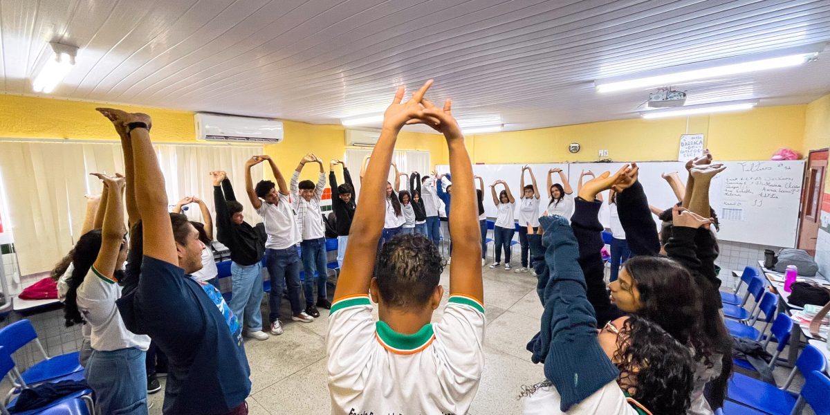 Na foto, o professor Vinícius e os alunos estão com os braços erguidos, se alongando durante a oficina de percussão corporal.